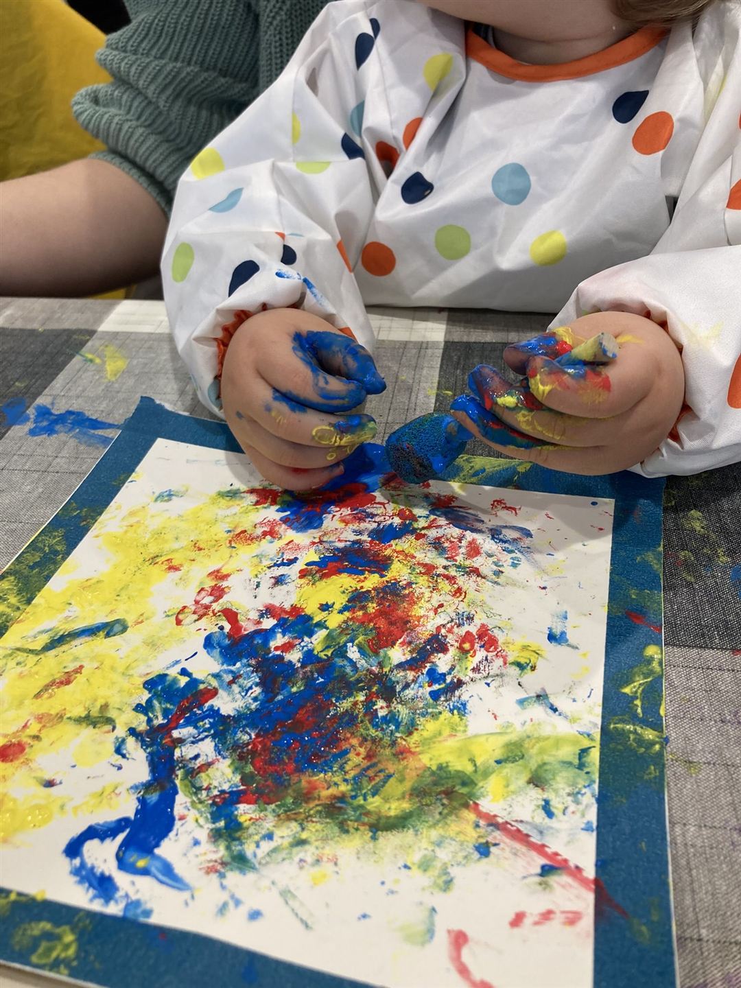 Bunte Hände eines Kleinkindes halten einen Schwammpinsel. Auf dem Tisch davor liegt ein bemaltes Papier.