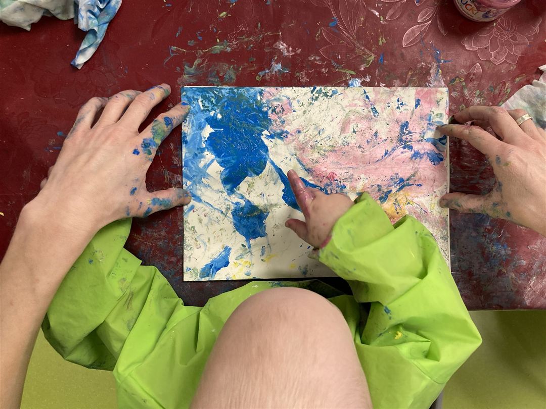 Hand eines Kleinkindes malt mit Fingermalfarbe ein Bild. Links und rechts des Papiers halten erwachsene Hände dieses fest.