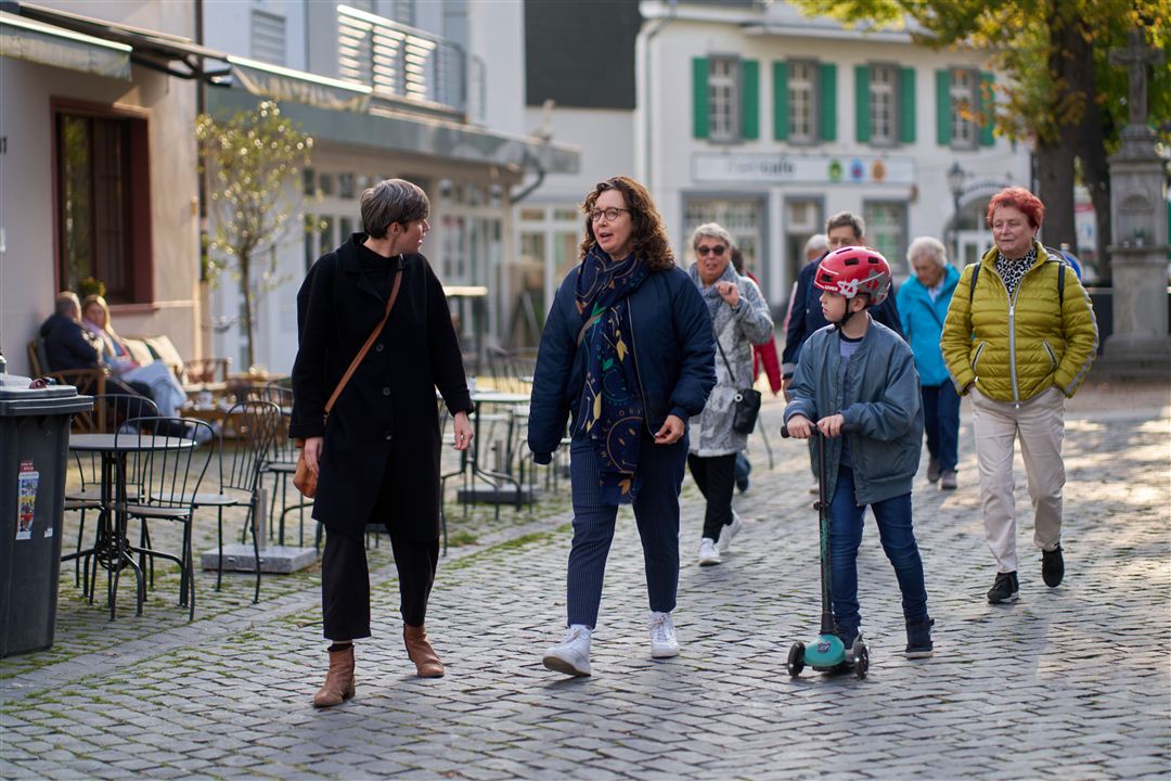 Mehrere Personen gehen auf einem gepflasterten Weg in einer belebten Fußgängerzone mit Außengastronomie und historischen Gebäuden.