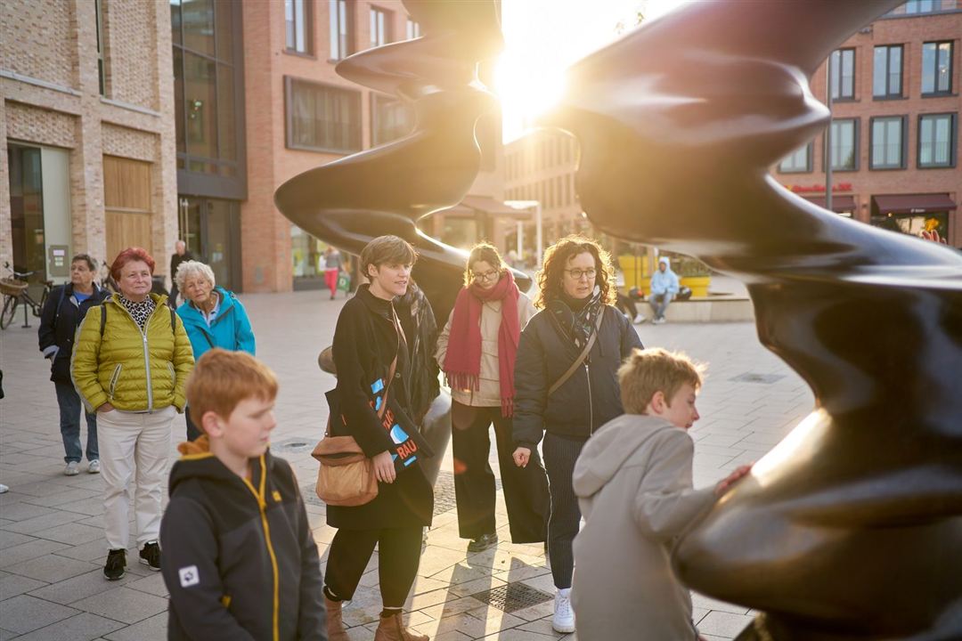 Mehrere Personen stehen um eine große, schwarze, spiralförmige Skulptur auf einem Platz mit Backsteinbauten im Hintergrund, die Sonne scheint von hinten.