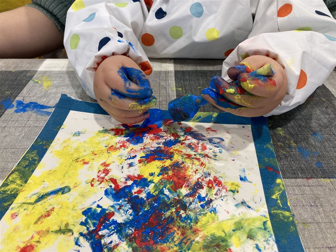 Kinderh&auml;nde mit bunter Fingerfarbe &uuml;ber einem Papier mit gelben, roten und blauen Farbflecken