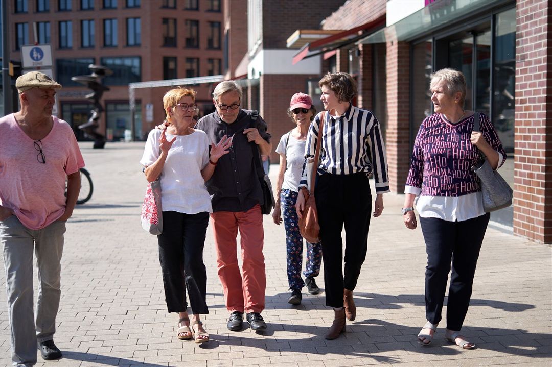 Gruppe von sechs Personen, die auf einem gepflasterten Platz vor einem modernen Gebäude gehen, darunter eine Frau mit gestreiftem Hemd und eine mit weißem Oberteil.