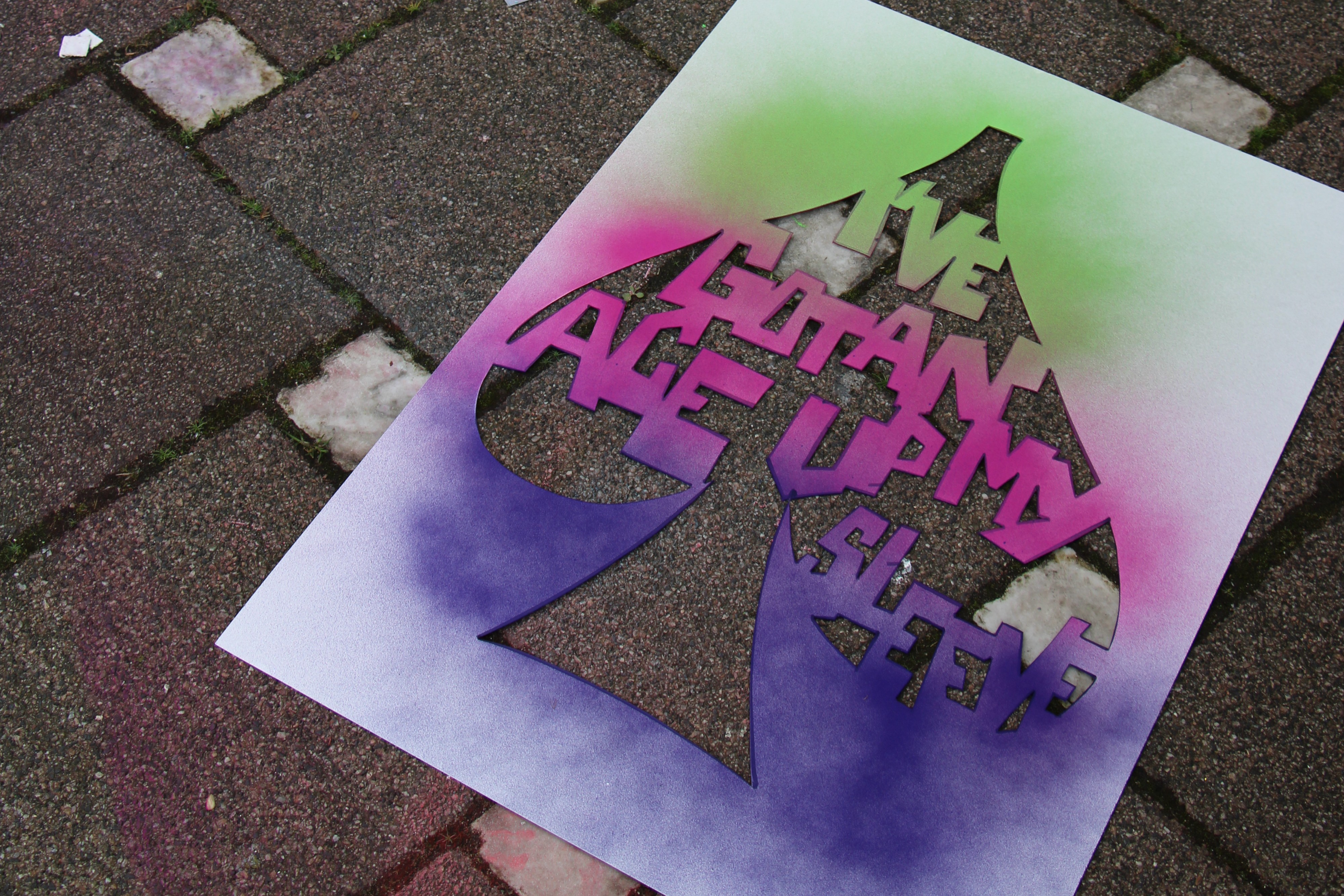 Eine Schablone mit einem Schriftzug auf einem Straßenpflaster, die schon bunt angesprüht ist.