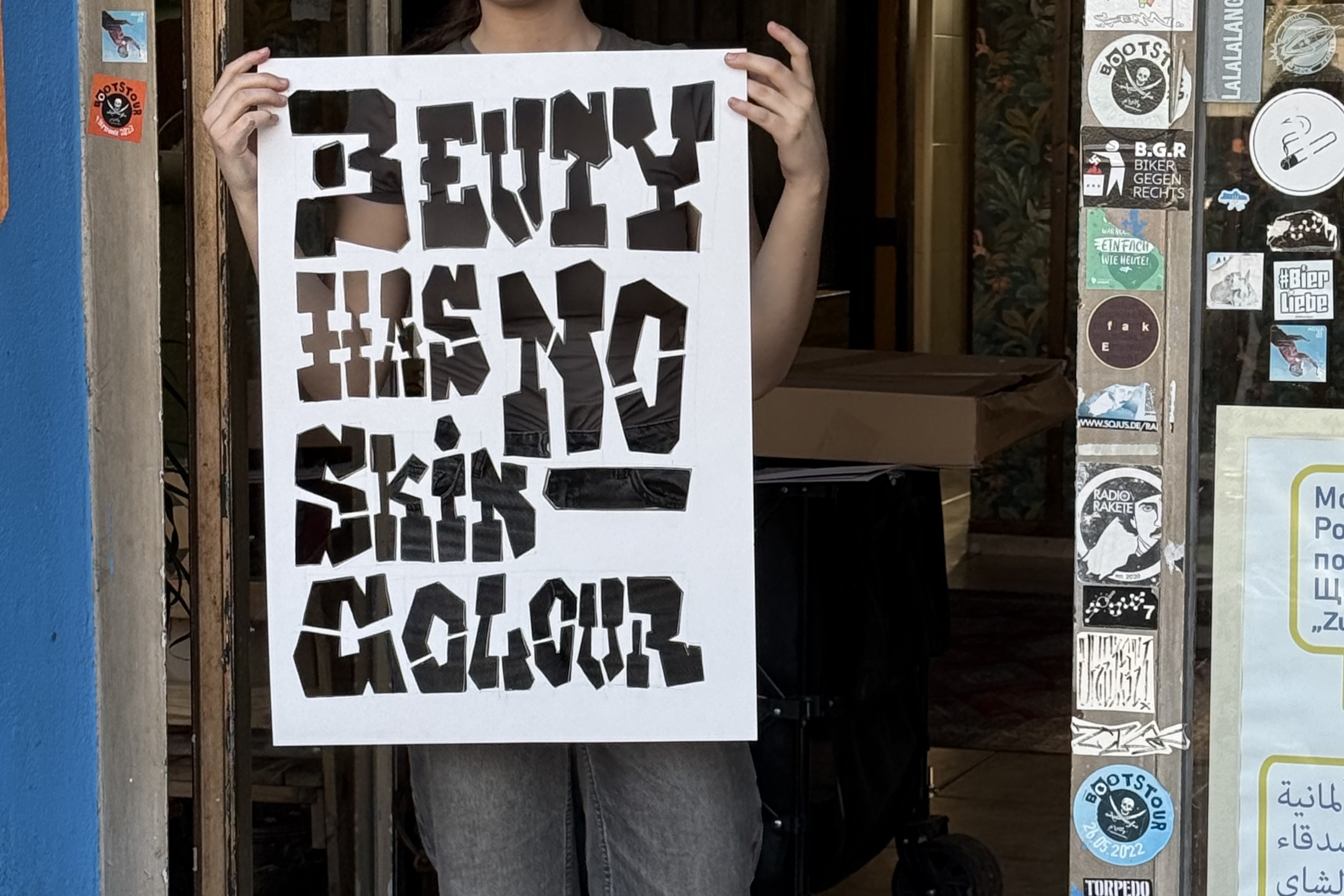Eine Person, die im Türrahmen eines Ladens steht. Sie hält eine Schablone mit dem Schriftzug "Beauty has no skin colour" hoch.