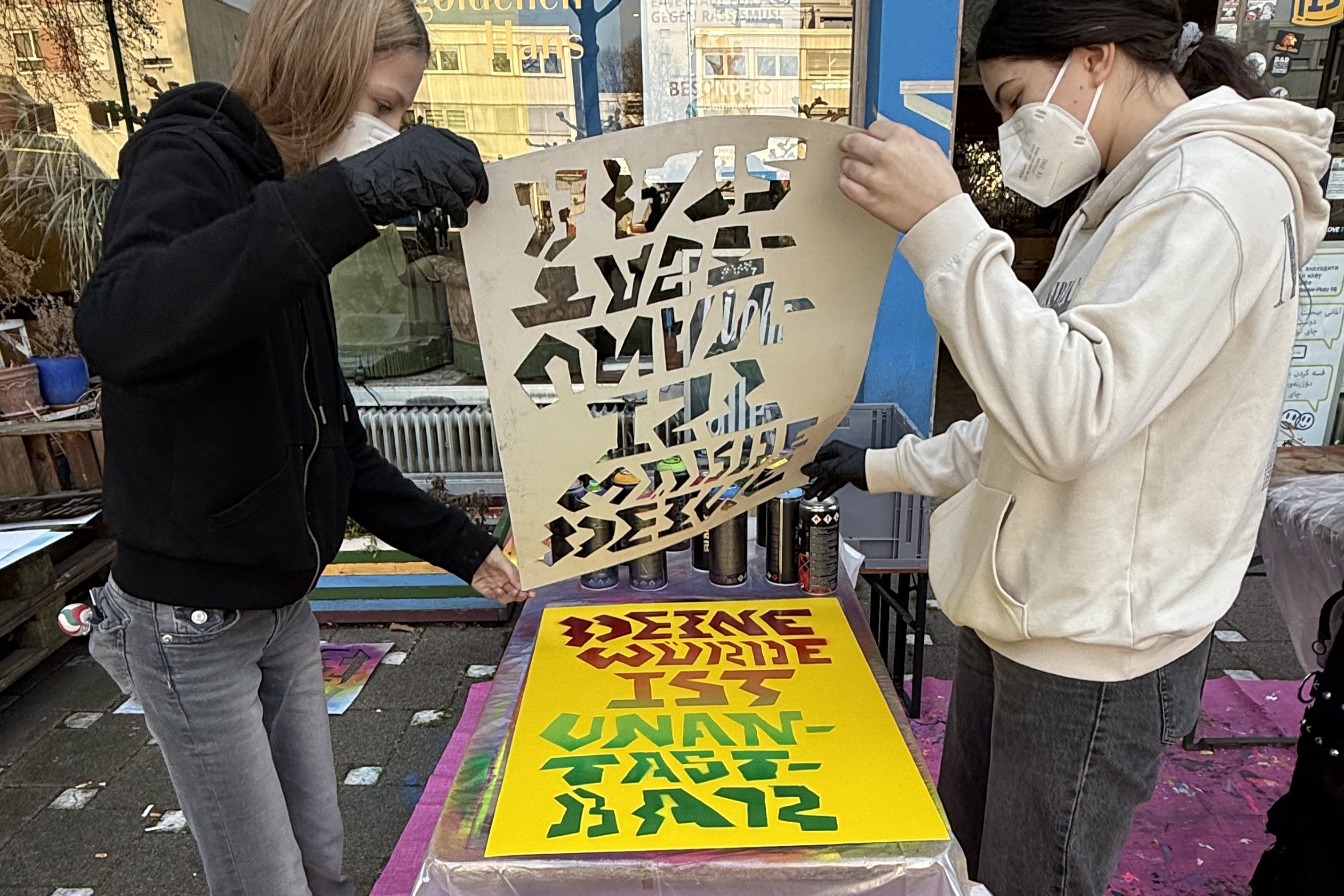 Zwei Personen heben eine Schablone von einem gelben Blatt Papier hoch, auf das sie gerade in bunt einen Schriftzug gesprüht haben.