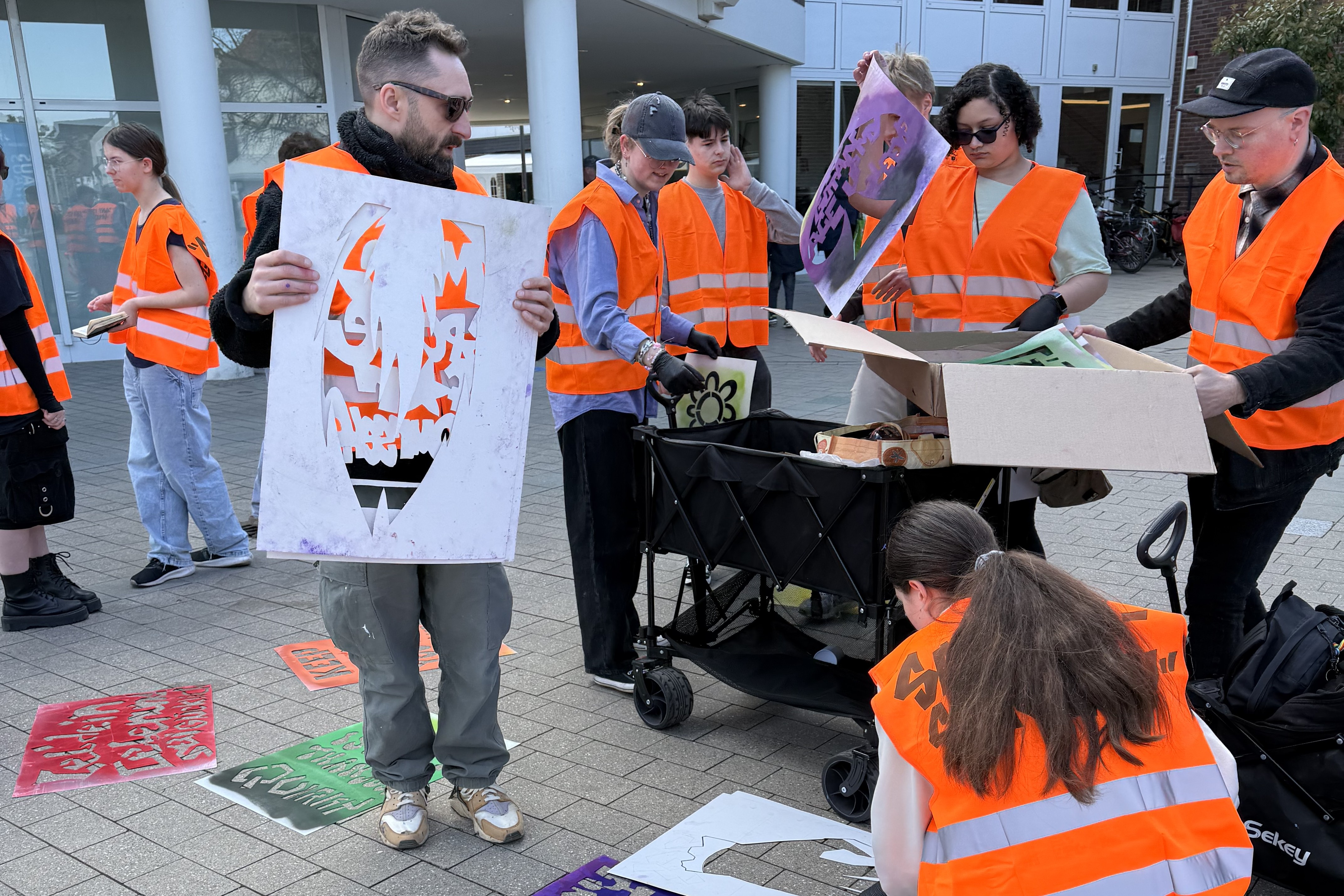 Mehrere Personen in orangenen Warnwesten im öffentlichen Stadtraum. Eine Person hält eine Schablone hoch, auf dem Boden sind verschiedene Graffitis.