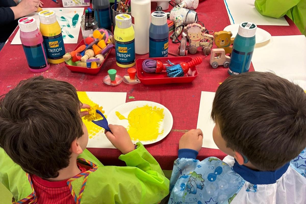 Blick von hinten über die Schultern zweier Kinder, die an einem Tisch mit Farben und Materialien sitzen.