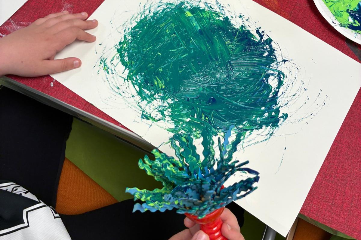 Auf einem Tisch liegt ein Papier, auf dem türkis-blaue Farbe ist. Im Vordergrund hält eine Hand einen großen Stempel, mit dem die Farbe auf dem Papier zuvor bearbeitet wurde.